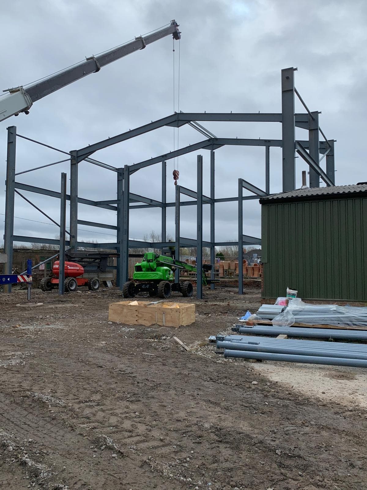 Crane-assisted erection of a large multi-bay steel portal frame alongside existing green-clad industrial buildings